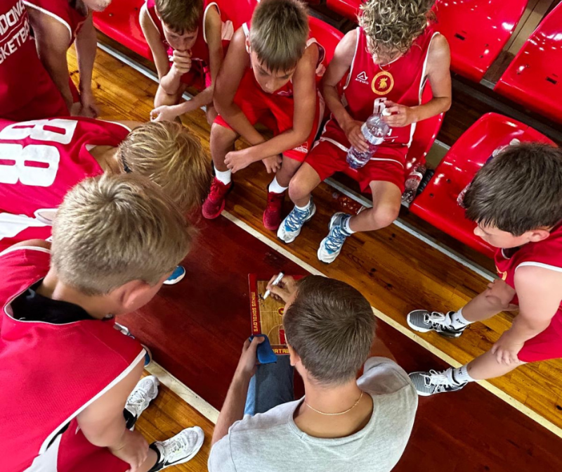 Mikus Sokolovs zīmē uz basketbola laukuma tāfeles, skaidrojot puišu komandai spēles taktiku/shēmas