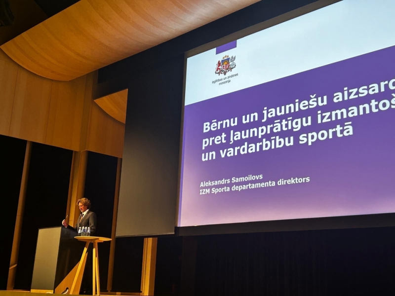 IZM Sporta departamenta direktors Aleksands Samoilovs konferencē stāv uz skatuves. Aiz viņa liels ekrāns ar prezentāciju uz violeta fona.