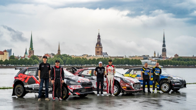 Fonā Rīgas ainava, priekšplānā 3 rallija mašīnas un pie katras stāv 2 sportisti