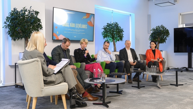 Izglītības un zinātnes ministre Anda Čakša piedalās diskusijā; septiņi cilvēki sēž pusaplī, ministre runā mikrofonā