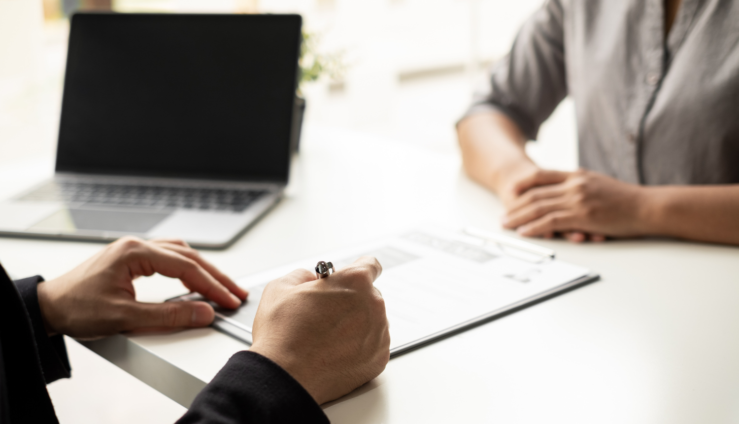 Uz galda stāv dators. Divi cilvēki sēž pretī viens otram, viens no cilvēkiem veic pierakstus.