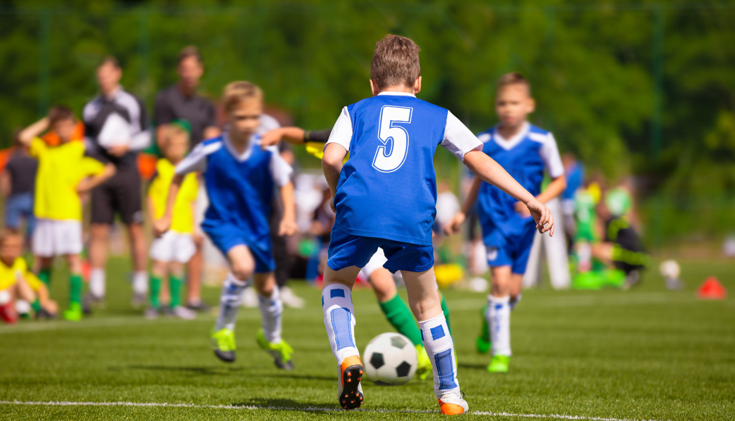 Puikas zilās formās spēlē futbolu. Fonā stāv citi puikas dzeltenās formās un treneri.