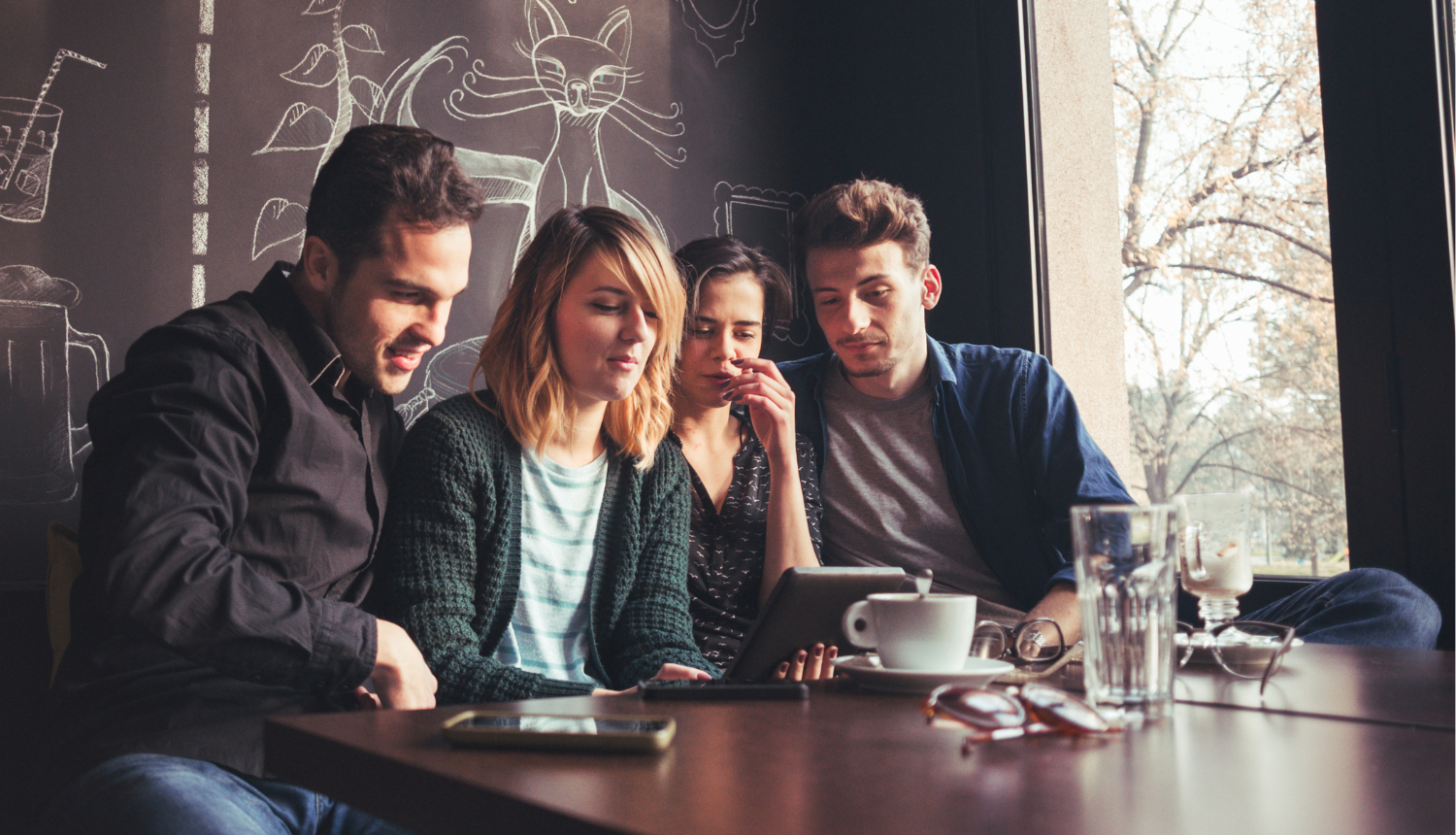 Četri jaunieši sēž kafejnīcā un sarunājas videozvanā caur planšetdatoru