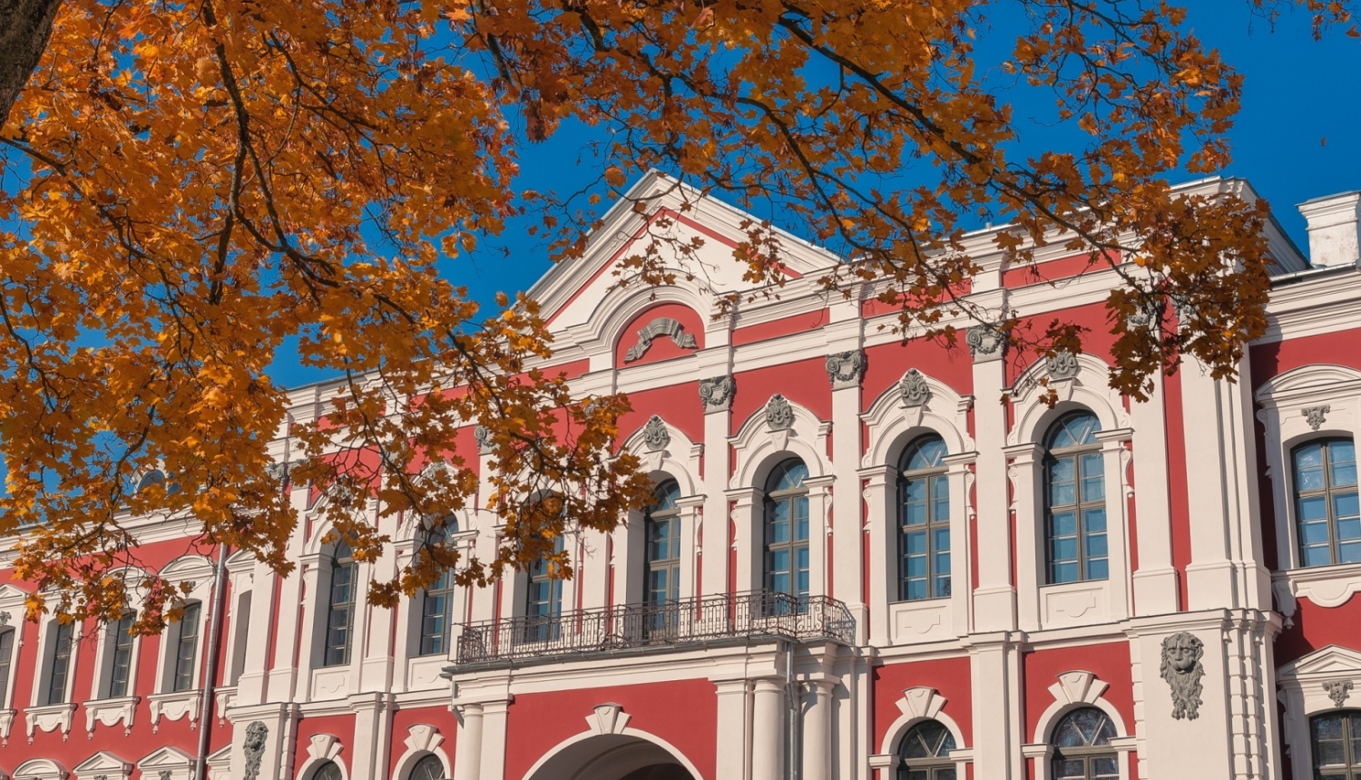 Latvijas Biozinātņu un tehnoloģiju universitātes ēka - klasiskā stila pils sarkanā krāsā ar baltiem logu rāmjiem un kolonnām. Priekšplānā redzam nelieks klavas zars ar rudenīgām, dzeltenām lapām